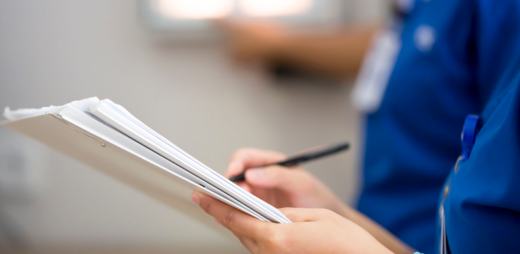 Close-up of a healthcare worker in blue scrubs writing on a clipboard