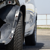 Damaged white car with a missing bumper and exposed wiring, parked near a fuel tanker, suggesting a recent accident