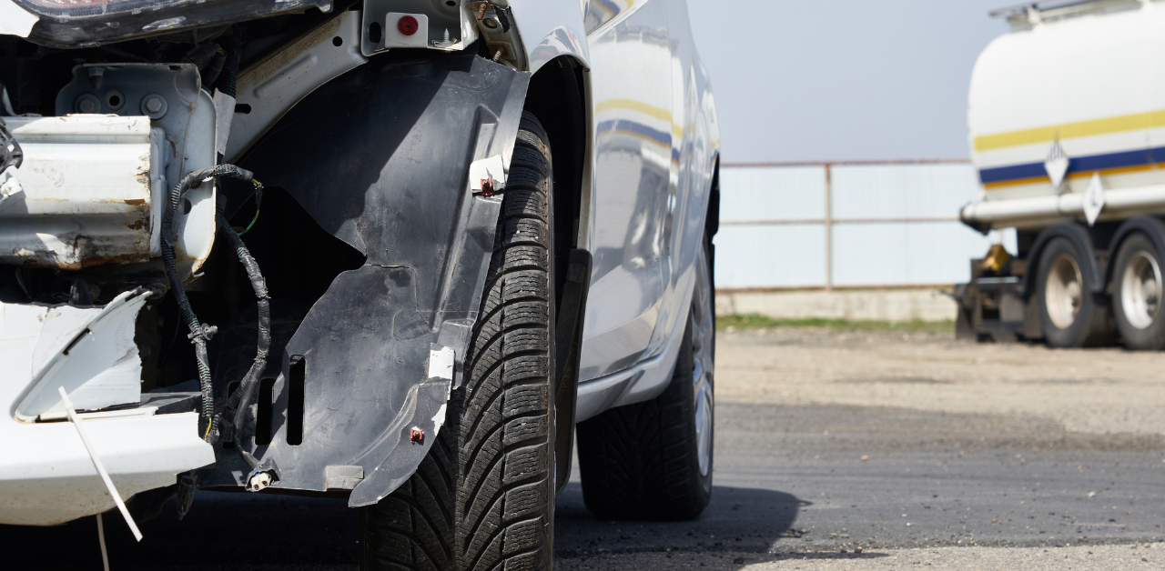Damaged white car with a missing bumper and exposed wiring, parked near a fuel tanker, suggesting a recent accident