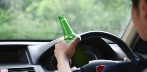 Driver holding a green beer bottle while gripping the steering wheel, symbolizing drunk driving danger