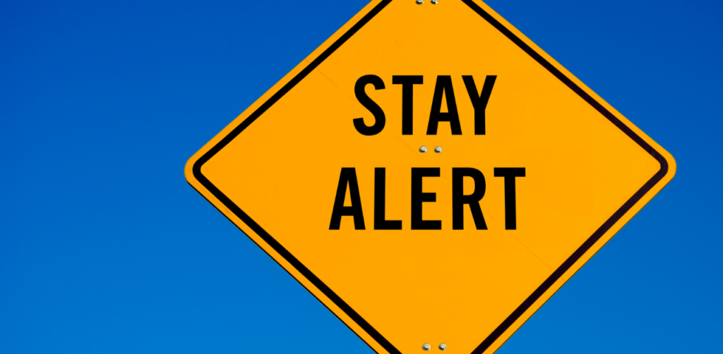 Yellow 'Stay Alert' road sign against a clear blue sky, warning drivers to be cautious