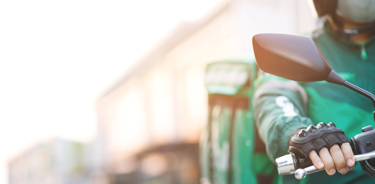Delivery rider in a green uniform grips a motorbike handlebar with a blurred urban street in the background