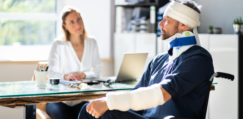 Injured man with bandages and neck brace sits in a wheelchair speaking to a professional woman at a desk, possibly a legal or medical consultation