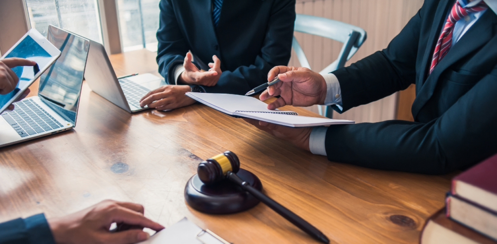 Lawyers discuss paperwork at a meeting table with laptops and a gavel, reviewing a legal settlement