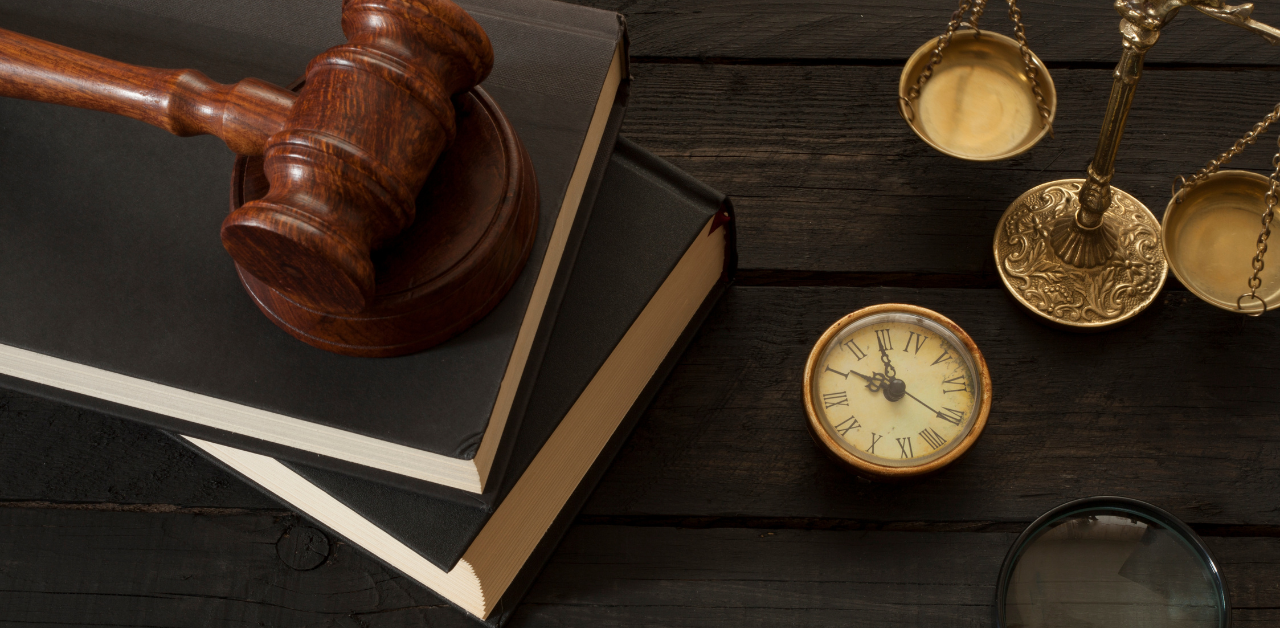 Wooden gavel on law books next to antique scales of justice and a vintage clock on a dark wooden table