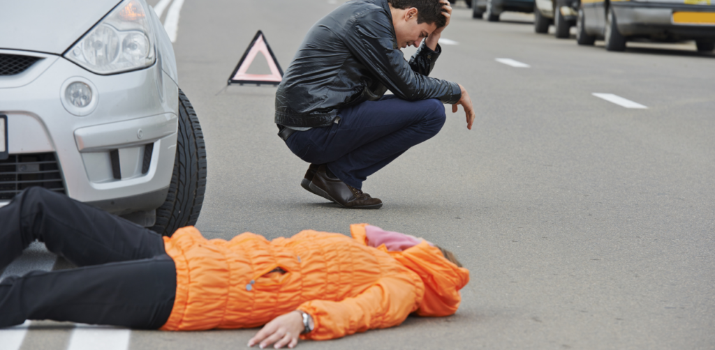 Distressed man crouching next to a person lying unconscious in the street after a pedestrian accident, with a warning triangle placed behind a silver car