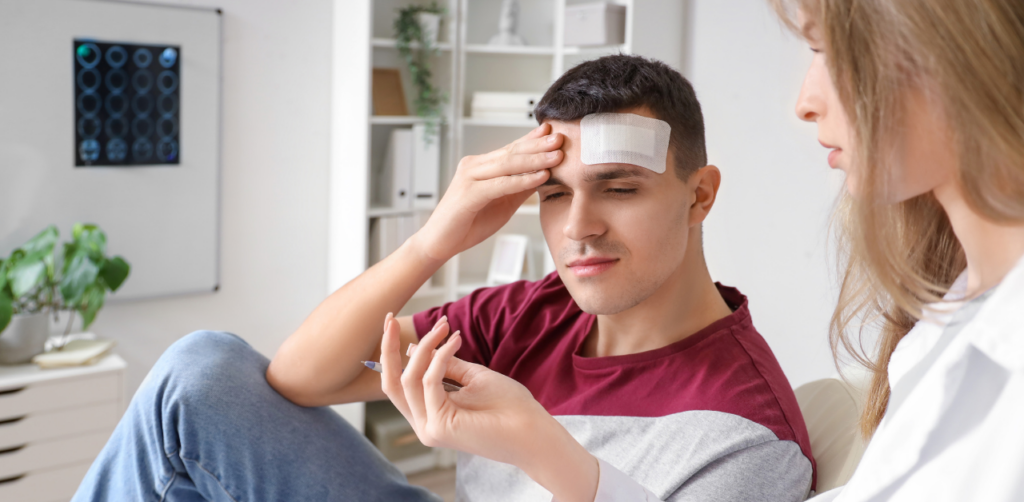 Injured man with head bandage holding his forehead while speaking to a female doctor in a medical office with brain scans in the background, illustrating a potential traumatic brain injury