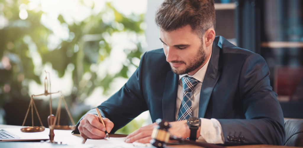 Male attorney in a suit writing on legal documents at a desk with a gavel and scales of justice nearby