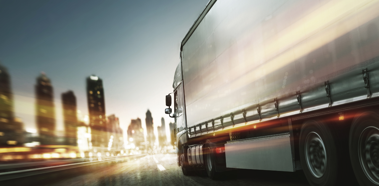 Motion-blurred semi-truck driving through a city at dusk, with illuminated skyscrapers in the background