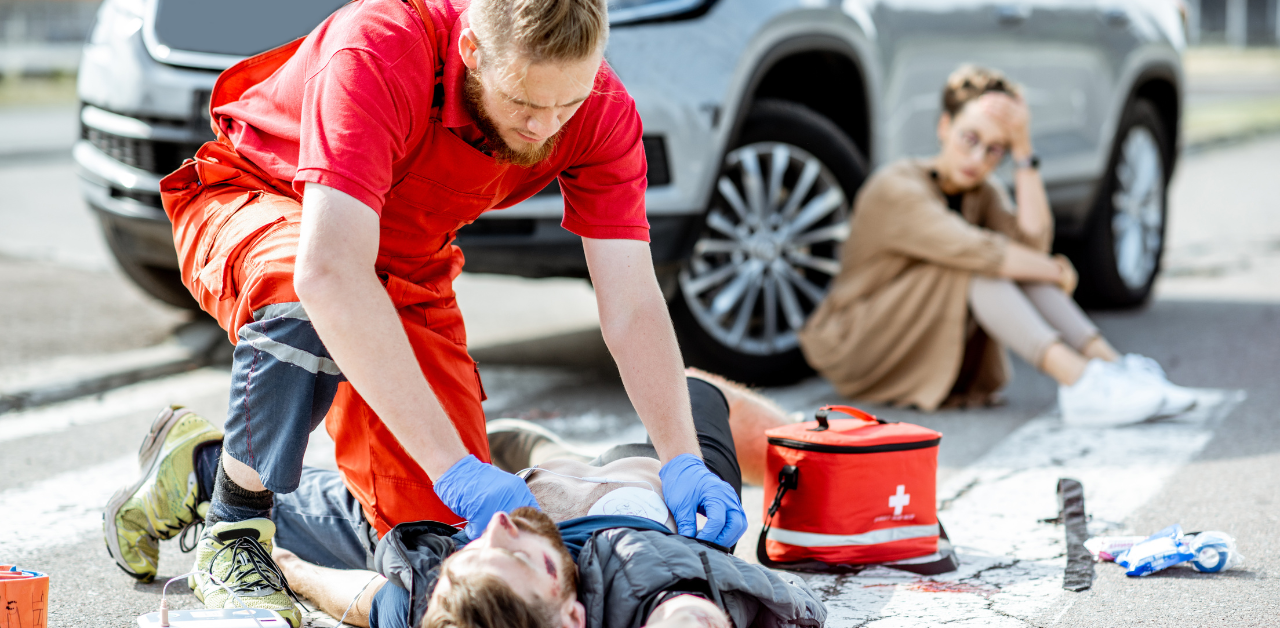 Paramedic performs CPR on man after car accident, while a distressed woman sits nearby on the road