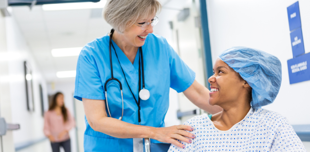 Nurse smiles at patient in wheelchair wearing hospital gown and cap in medical hallway