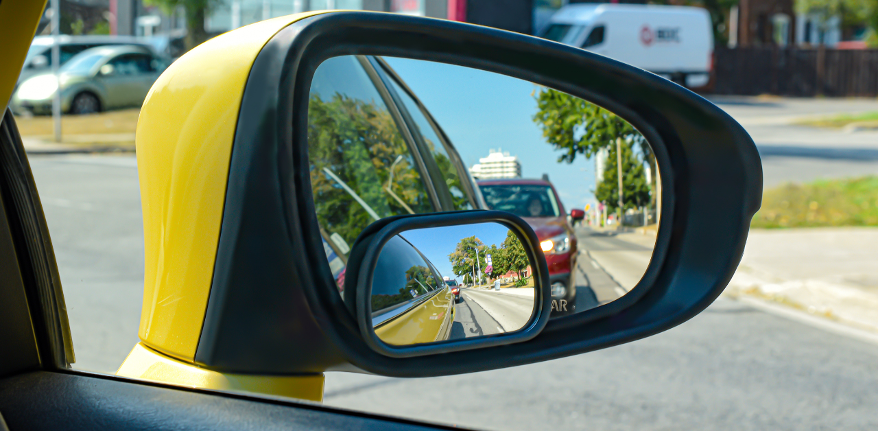 Rearview mirror of a yellow car showing trailing traffic and trees, captured by a vehicle camera system