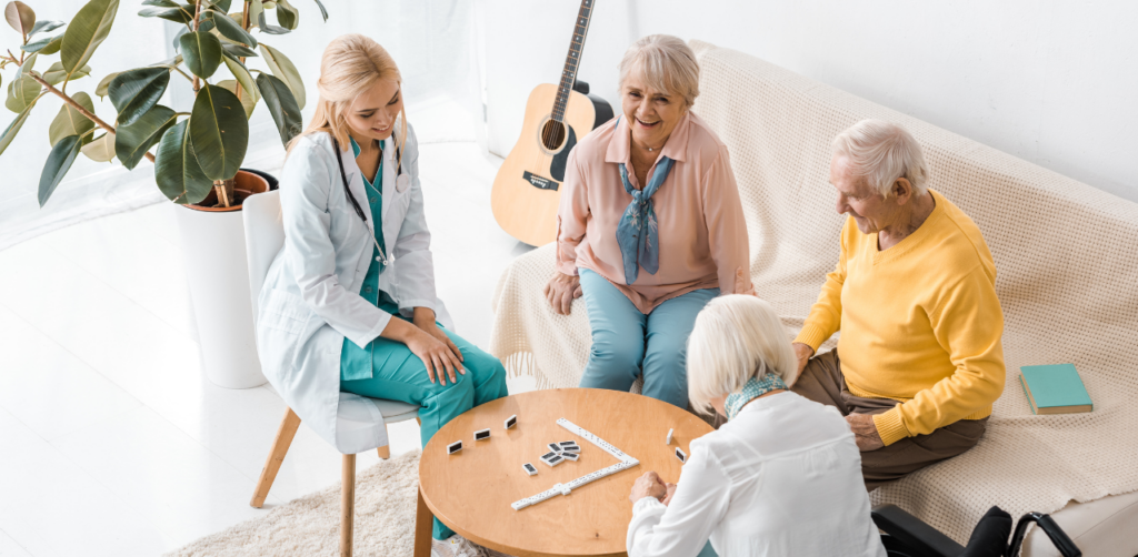 Smiling seniors play dominoes with a healthcare worker in a bright room with a guitar and houseplants