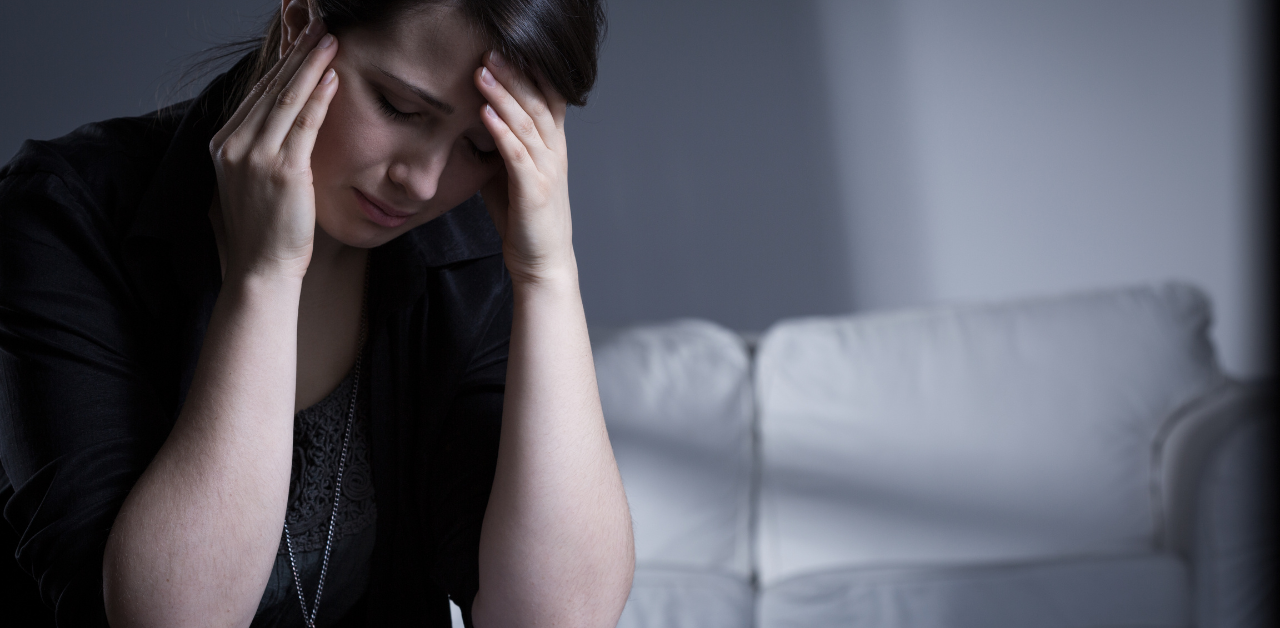 Stressed woman sits alone on couch in dim room, holding head in hands with visible anxiety