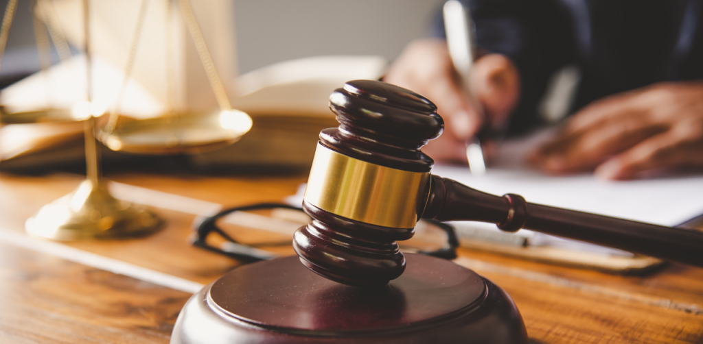 Wooden gavel and golden scales of justice on a desk with a person writing in the background, symbolizing a legal case