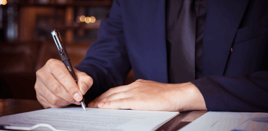A man in a navy suit signs legal documents at a desk, possibly related to a car accident claim