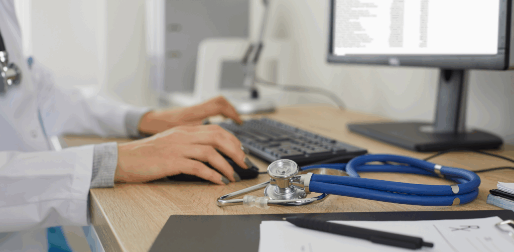 A physician types on a computer keyboard beside a stethoscope and medical documents on a desk