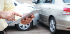 Man using a tablet to document a car accident with two damaged vehicles in the background
