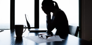 Silhouette of a woman sitting at a desk with her head in her hands in front of a laptop, papers, and coffee cup, conveying stress or frustration