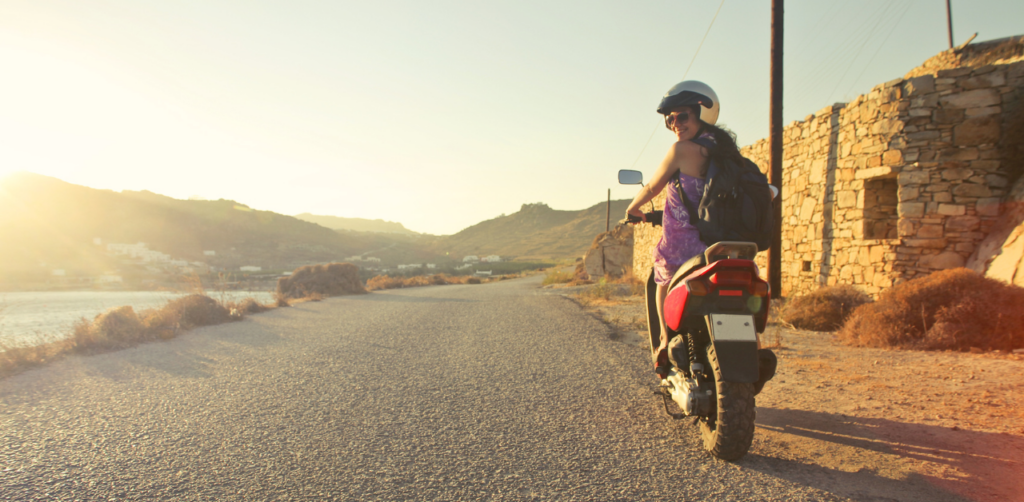 Smiling woman riding a scooter on a scenic coastal road at sunset