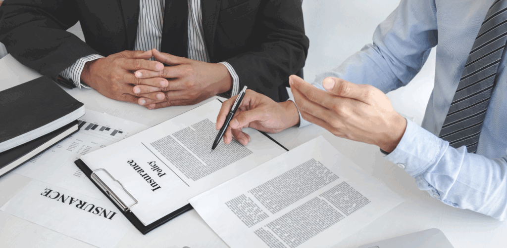 Two businessmen discuss an insurance policy document at a desk covered with papers and charts