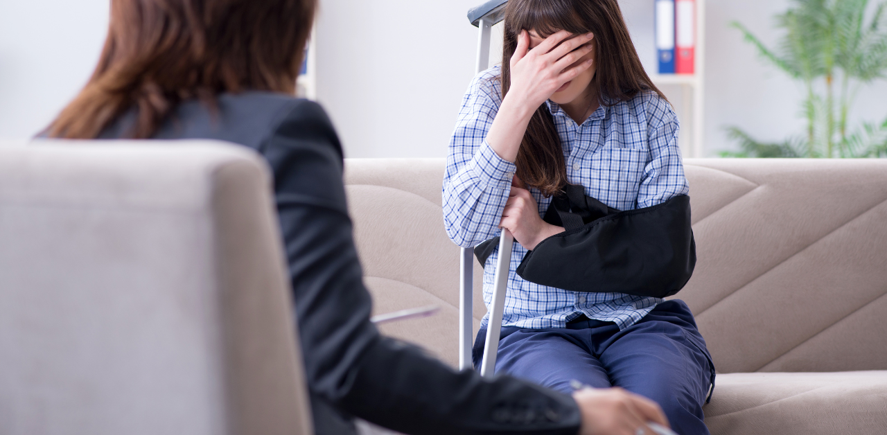 Distressed woman with her arm in a sling sits on a couch, covering her face while holding a crutch, speaking with a professional lawyer about her personal injury case
