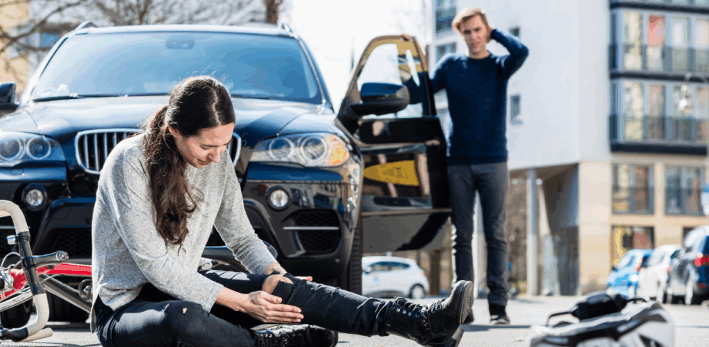 Female cyclist sits on the ground clutching her injured leg in front of a black SUV, with a concerned man standing by the open car door, depicting the aftermath of a vehicle-bicycle accident