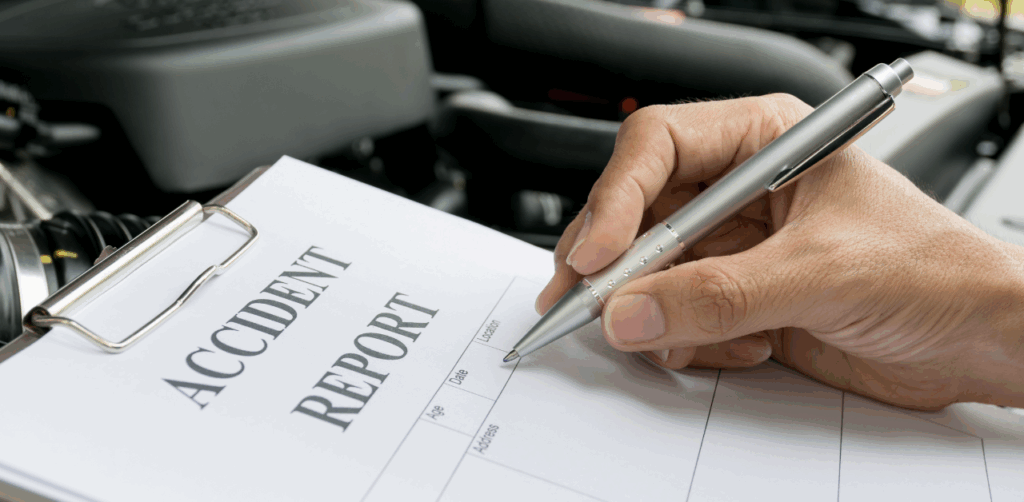 Hand fills out an “Accident Report” form on a clipboard beside an open car hood, documenting details for an insurance or legal case