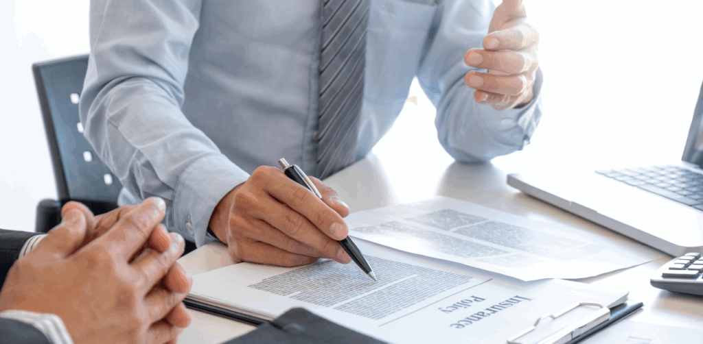 Man in a blue shirt and striped tie explains details of an insurance policy document to another person during a formal meeting, emphasizing the importance of understanding legal coverage