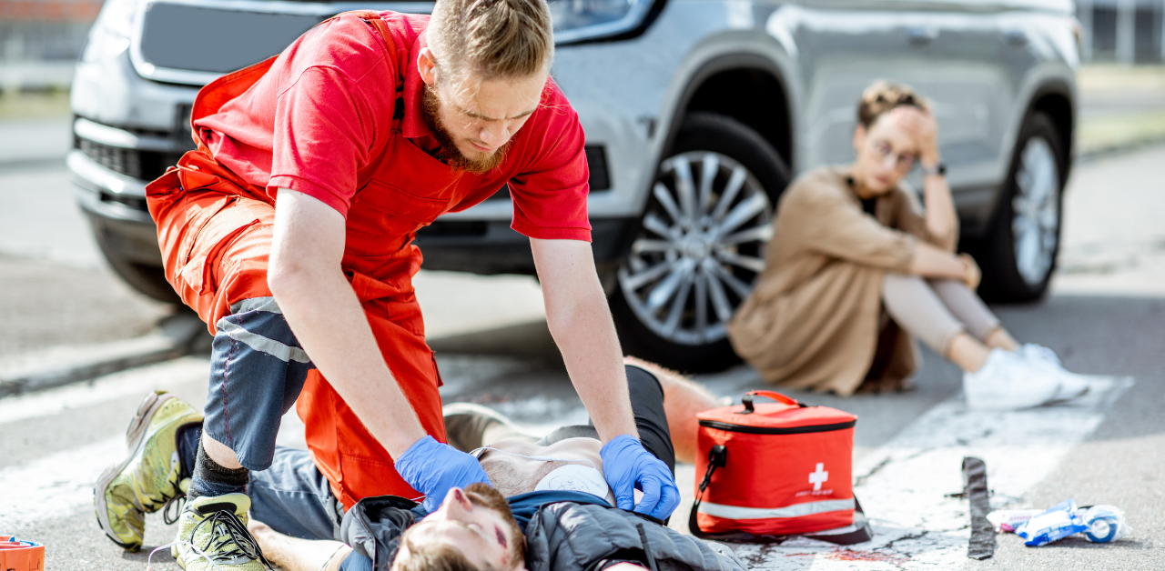 Paramedic in red uniform performs CPR on an unconscious man lying on a crosswalk, with a red first aid kit nearby and a distressed woman sitting by a silver SUV