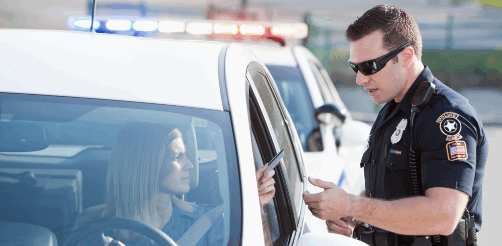 Police officer wearing sunglasses speaks to a woman in her car during a traffic stop, holding her license or ID while police lights flash in the background