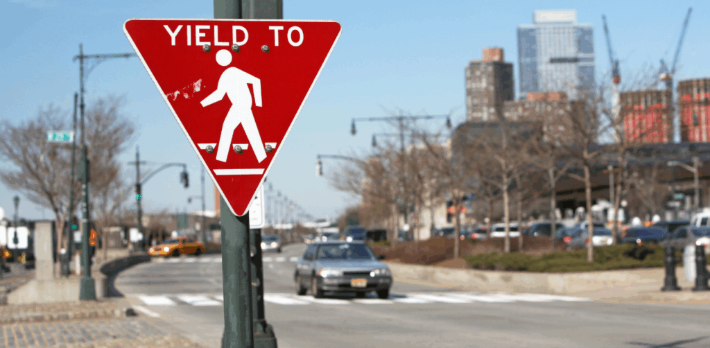 Red triangular “Yield to Pedestrians” sign is mounted on a city street pole, with blurred traffic and tall buildings in the background, promoting pedestrian safety and driver awareness