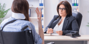 Serious-looking attorney listens as a woman in a neck brace and wheelchair explains her situation, depicting a personal injury consultation in a legal office