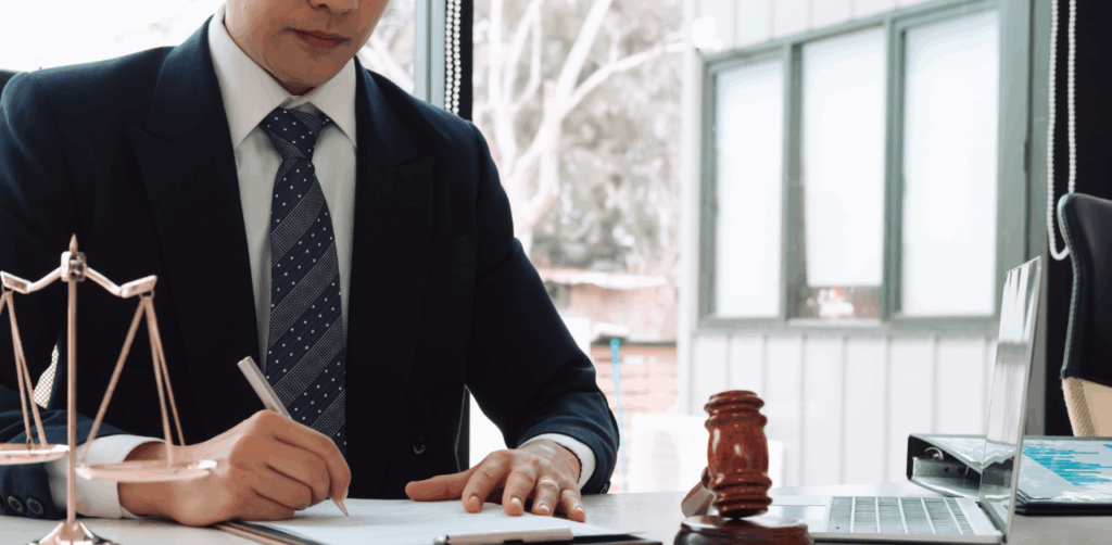 Lawyer in suit writing at desk with gavel, laptop, and justice scale near window-lit office