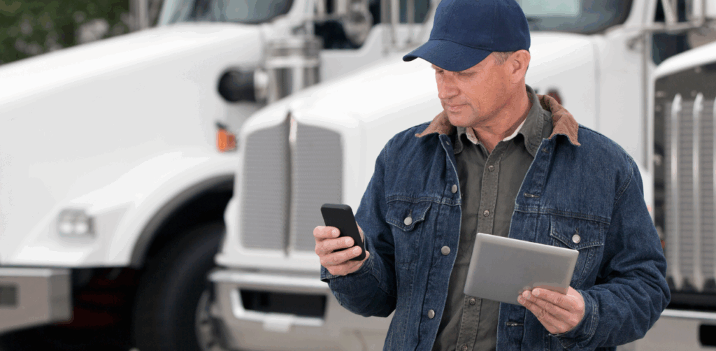 A truck driver stands in front of semi-trucks while using a smartphone and holding a tablet, possibly gathering evidence or communicating with a truck accident lawyer