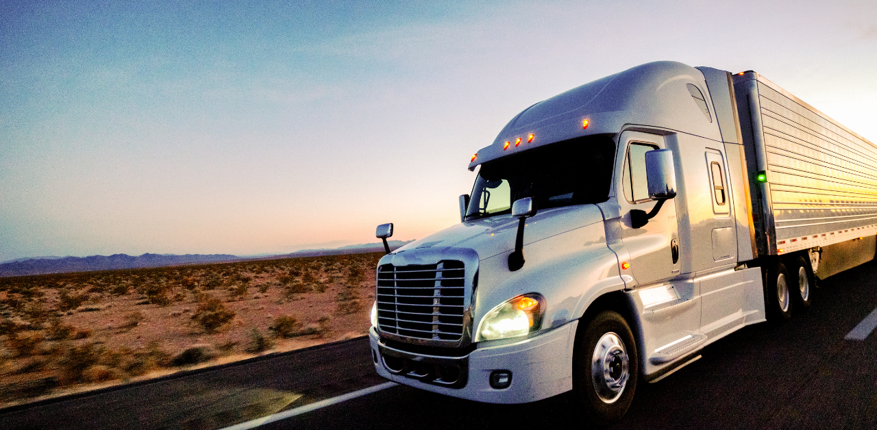 White semi-truck driving on a desert highway at sunset, headlights on and trailer reflecting light