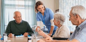 Caregiver helps seniors with medication at a table, showing warm senior medication assistance