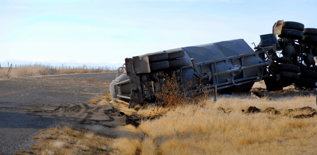 Overturned semi-truck lies off rural road, surrounded by dirt and dry grass
