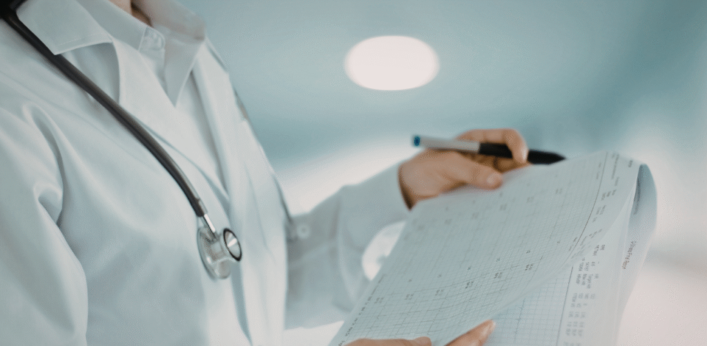 Doctor in white coat with stethoscope reviewing a medical chart and writing notes in a bright clinical setting