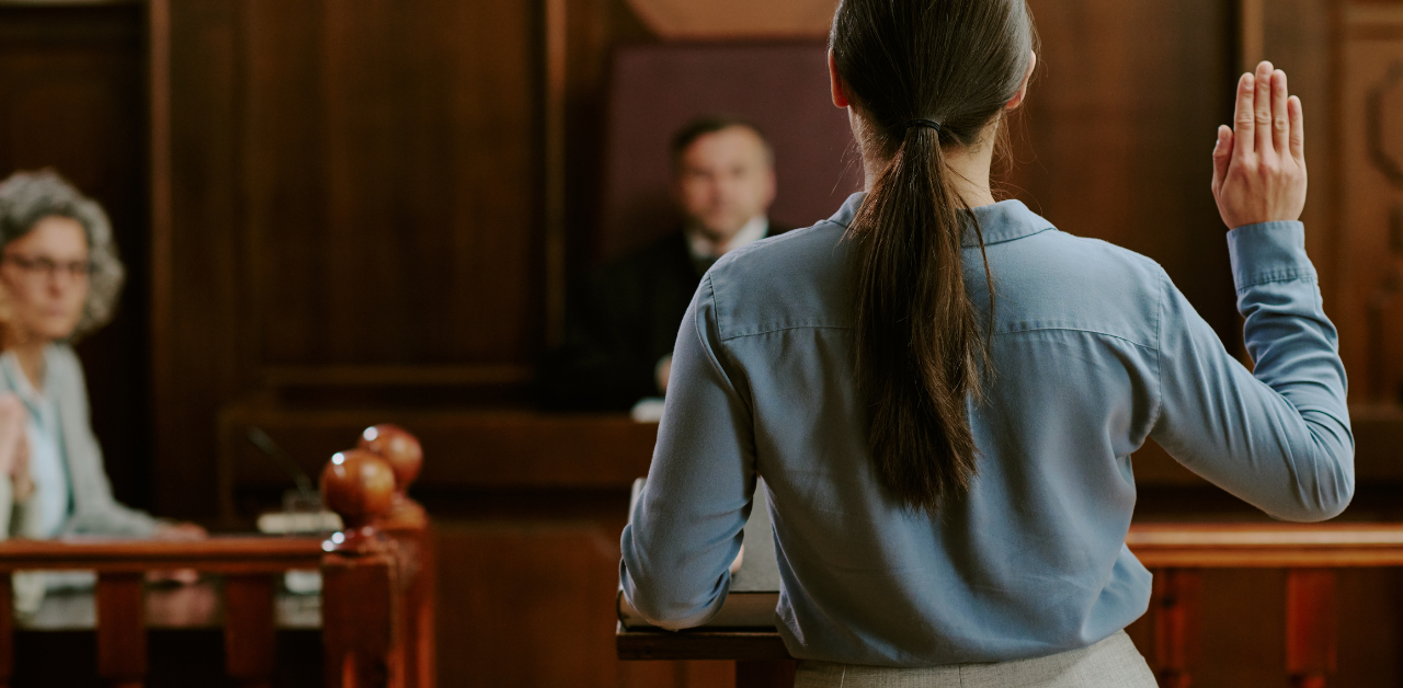 Woman testifying in courtroom with hand raised, judge and attorneys in background, highlighting legal trial proceedings