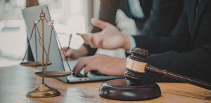 Close-up of a judge’s gavel and scales of justice on a desk while a legal professional works on a laptop in the background