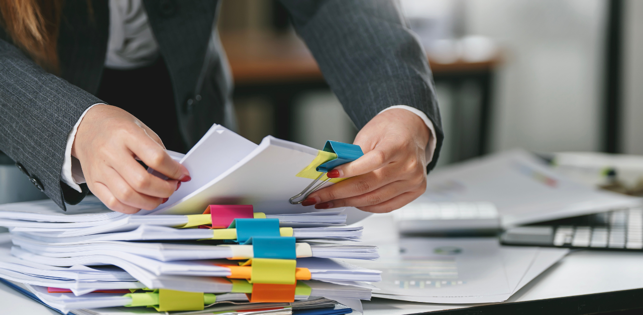 Person in a business suit organizing thick stacks of paperwork with colorful sticky tabs and binder clips on an office desk