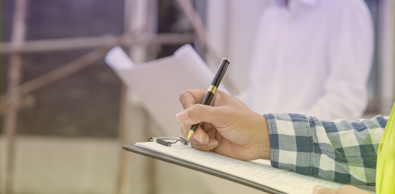 Person writing notes on a clipboard while inspecting or documenting information, with another person holding papers in the background
