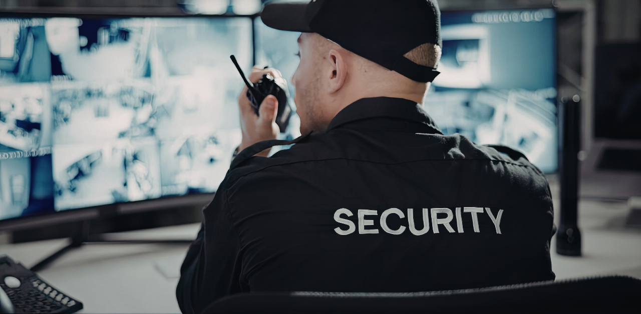 Security guard monitoring surveillance camera feeds on multiple screens while speaking into a handheld radio