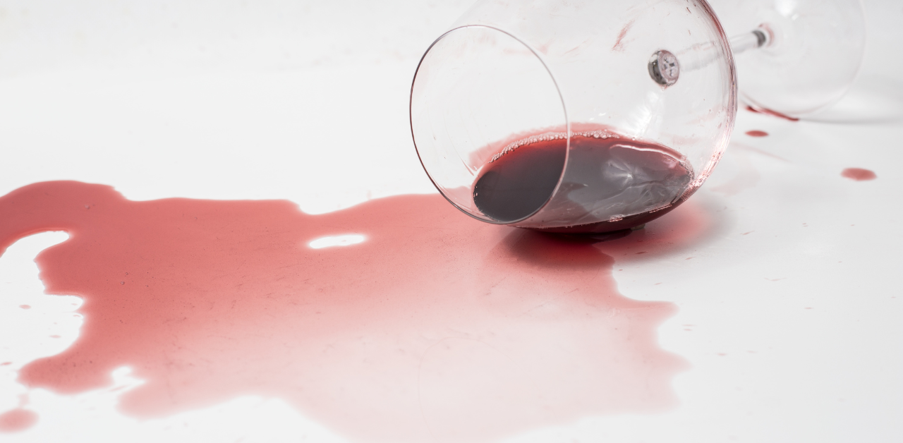 Spilled red wine from a tipped glass spreading across a white surface, creating a vivid liquid stain