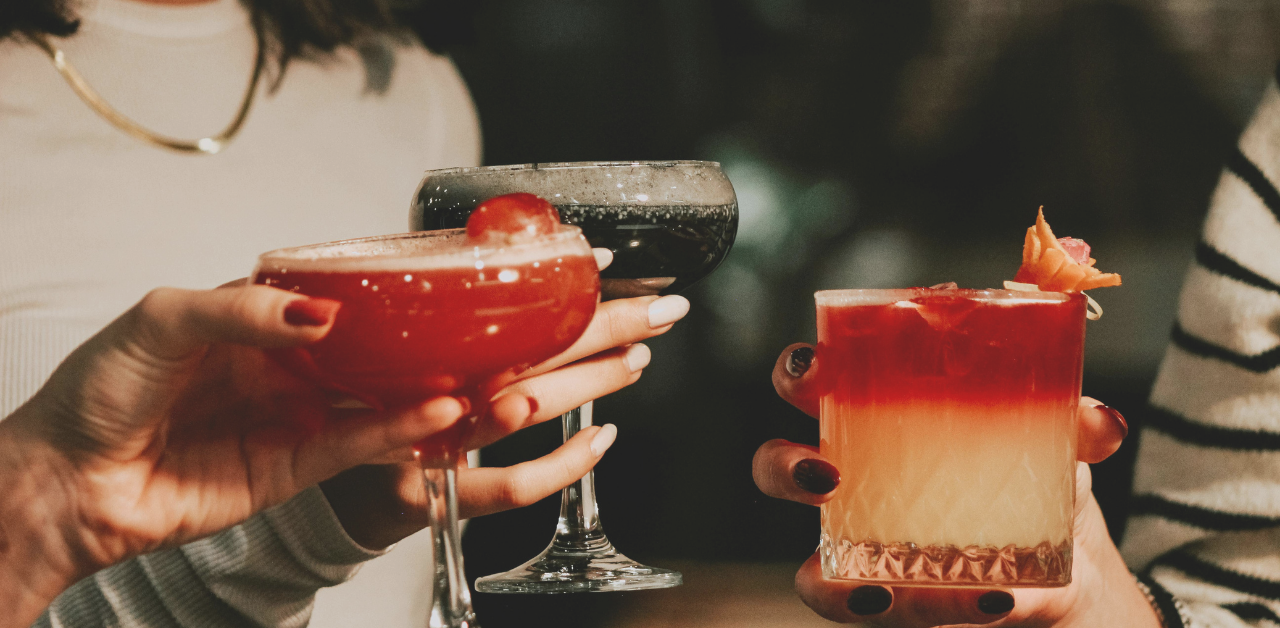Close-up of people holding colorful cocktails in a dimly lit social setting, with hands raised as if making a toast