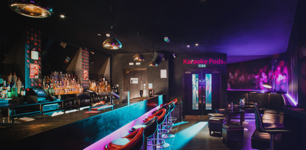 Interior of a dimly lit bar with neon lighting, seating areas, and a sign pointing to karaoke pods