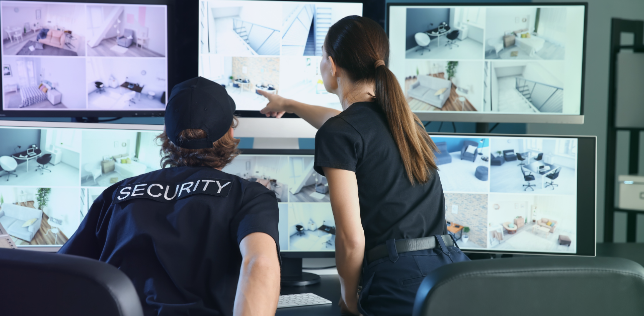 Two security staff members monitoring multiple surveillance screens in a control room, with one person pointing at footage displayed across several monitors