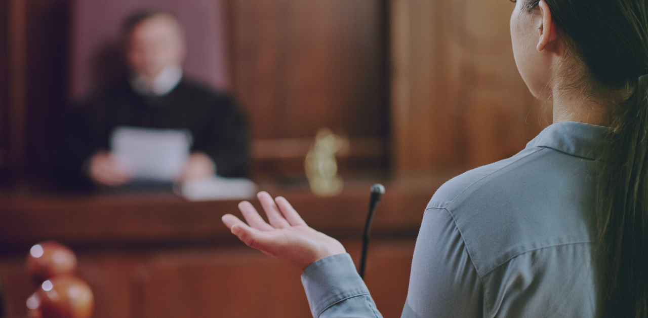 Person testifies from the witness stand while a judge listens from the bench in a courtroom