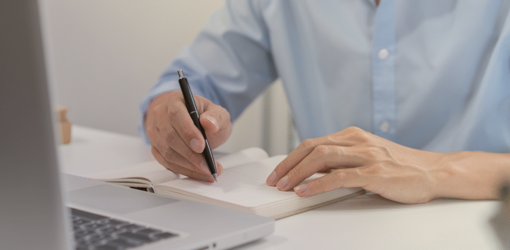 Person writes notes in a notebook beside an open laptop at a desk
